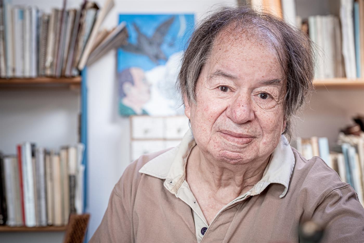 Homem idoso com camisa bege numa sala com livros e pintura ao fundo