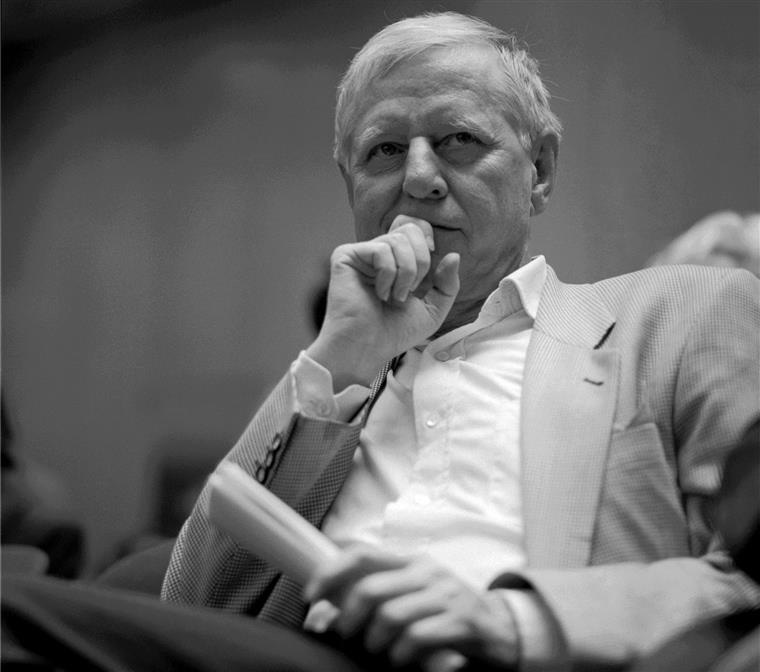 Homem idoso sentado com blusão de riscas e camisa branca