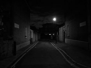 East Ending, Eduardo Brito Rua estreita à noite com edifícios de tijolo e iluminação de poste