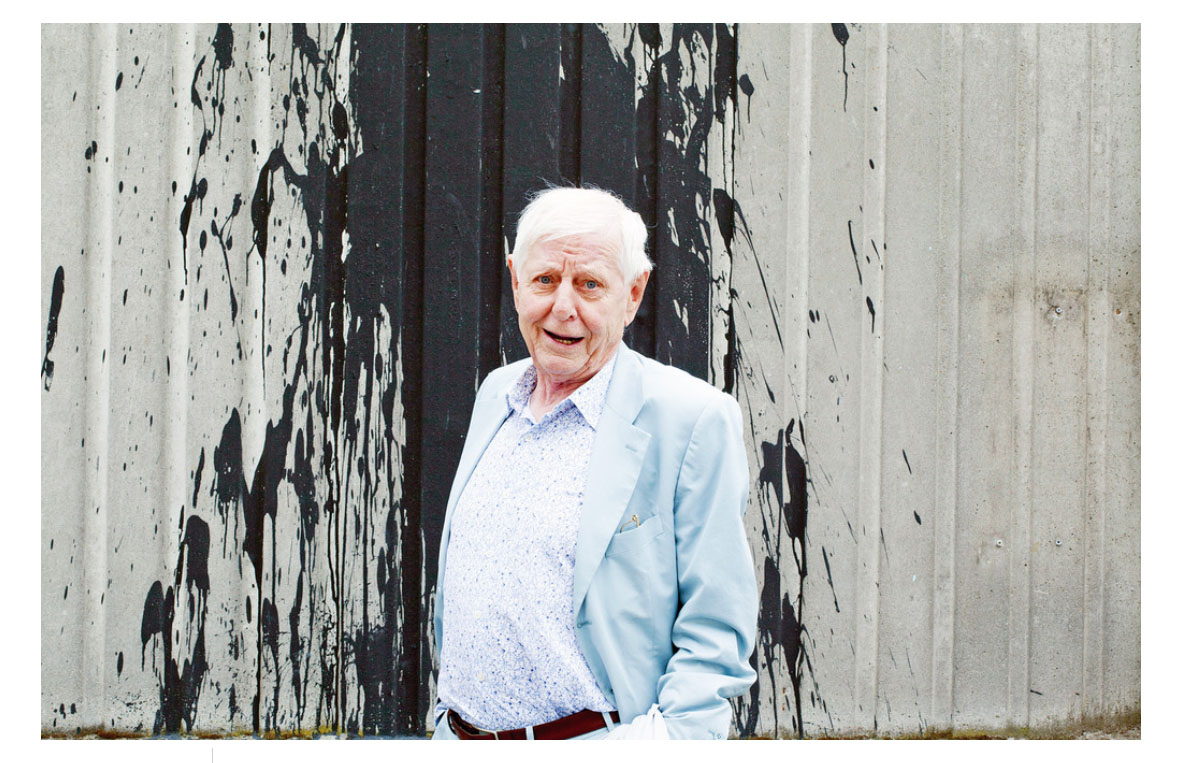 Homem idoso em roupa azul claro e branca junto a parede cinzenta com pintura preta escorrida