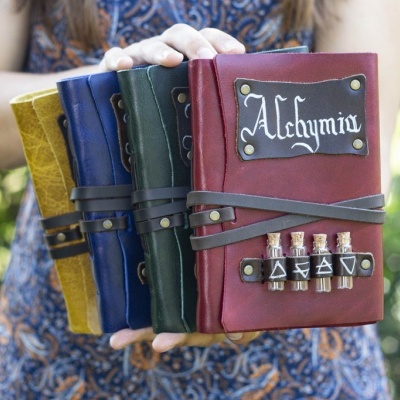 Quatro cadernos de capa dura coloridos com detalhes metálicos e texto 'Alchymia'