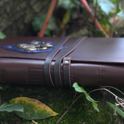 Caderno de pele castanha com fecho e distintivo metálico azul sobre musgo verde