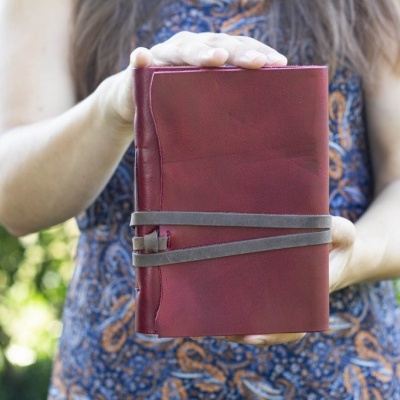 Caderno de couro vermelho fechado com fita castanha, mãos segurando, fundo com vegetação.