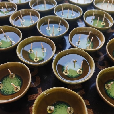 Velas em recipientes cerâmicos castanhos com interior verde escuro, com dois pavios, alinhadas numa superfície escura com letras marrons.