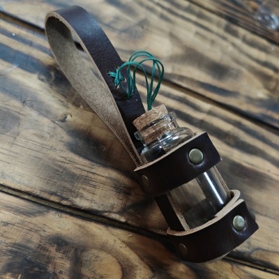 Frasco de vidro com rolha preso a cinta de couro castanho com alça, sobre madeira