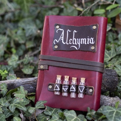 Caderno vermelho com placa 'Alchymia' e quatro frascos pequenos em couro sobre vegetação