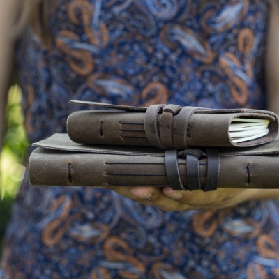 Duas agendas de couro castanho com tiras, seguradas por pessoa com vestido azul e padrão castanho, ao ar livre