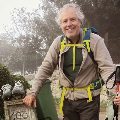 Homem com mochila azul e amarelo, casaco bege, junto a pedra com número 100 e ténis