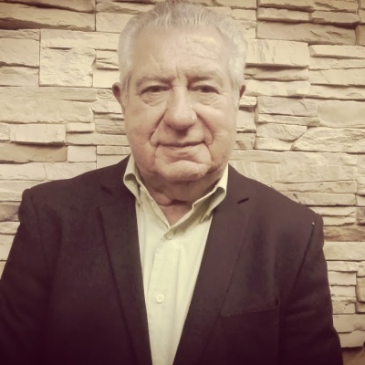 Homem idoso com camisa bege e blazer preto em frente a parede de pedras