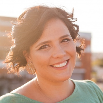 Mulher sorridente com brincos dourados e blusa verde em ambiente exterior