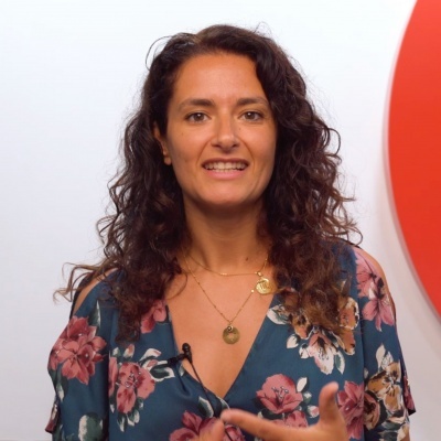 Mulher com blusa florida azul e colares dourados em fundo branco com círculo vermelho.