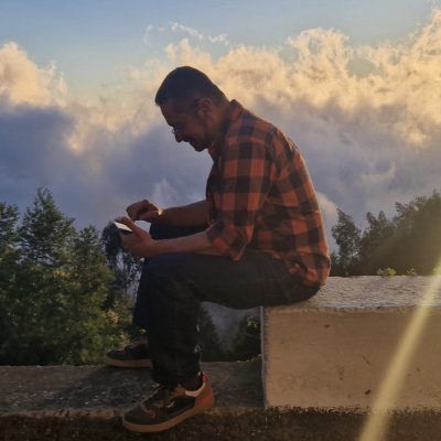 Homem sentado num muro com camisa xadrez laranja e preto usando telemóvel ao ar livre