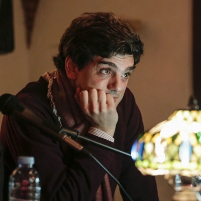 Homem sentado com casaco e cachecol junto a microfone e luminária colorida