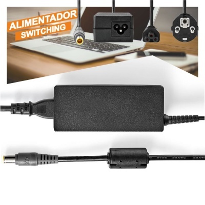 Alimentador switching preto com conector DC e três tipos de fichas de tomada em cima de uma mesa com computador portátil.