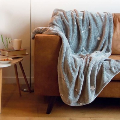 Manta cinza felpuda com nuvens sobre sofá castanho ao lado de mesa pequena com livros e caneca.