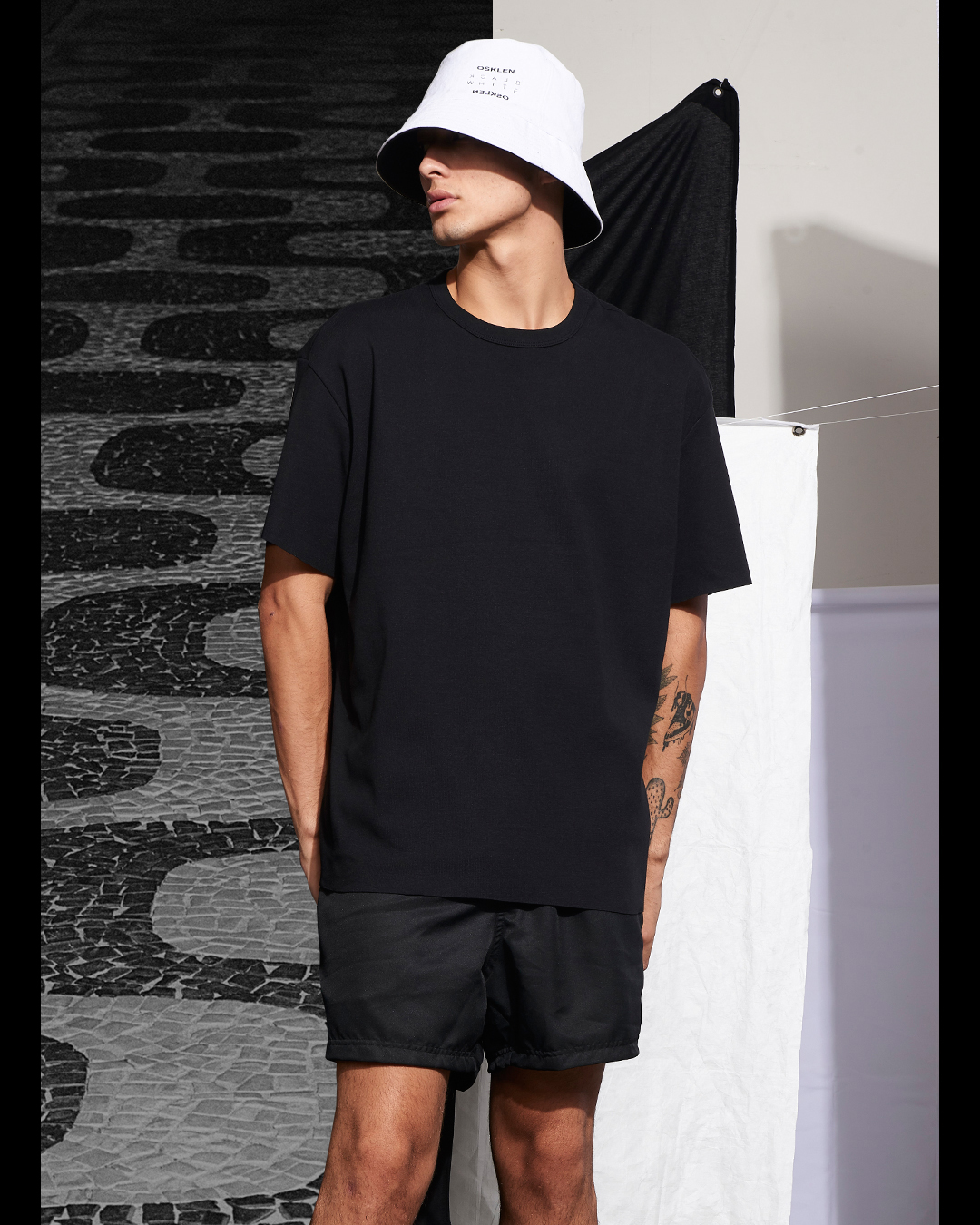 Homem com roupa casual preta e chapéu branco numa rua com pavimento de pedra mosaico