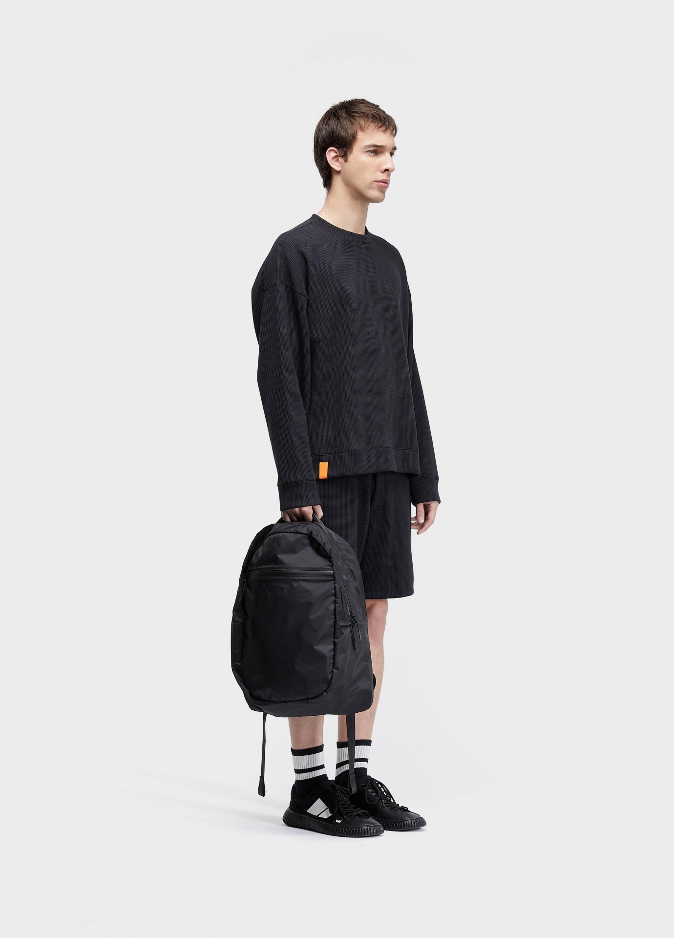 Bermuda Waffle Texture Osklen Homem com sweat preta e calções, meias brancas com riscas pretas e ténis na mão mochila preta