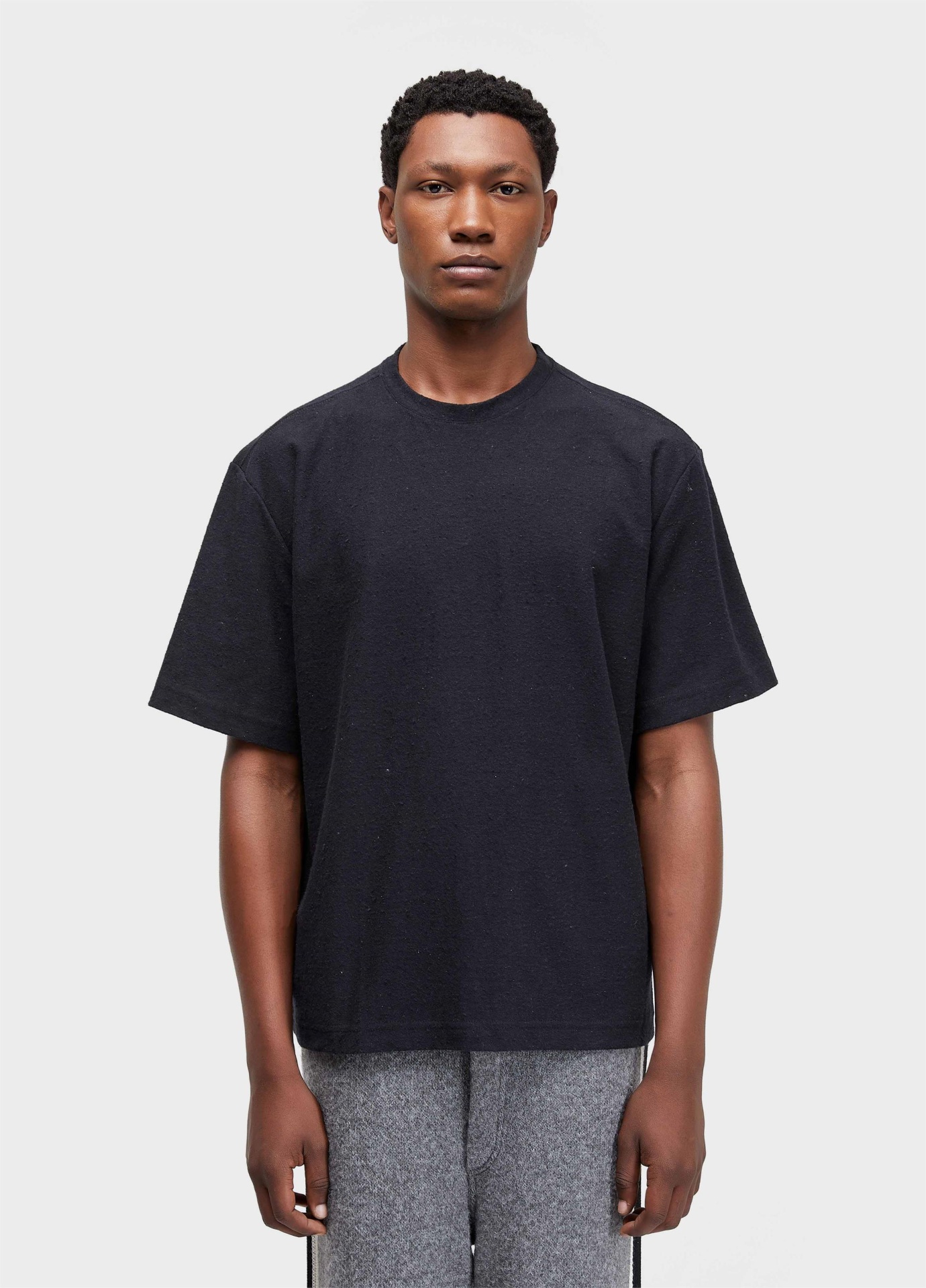 T-shirt Over Eco Osklen Homem de pele escura com t-shirt preta e calças cinzentas em fundo branco.