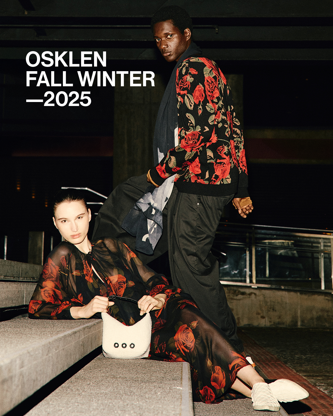 Dois modelos com roupas pretas com padrão de rosas vermelhas, ao ar livre à noite.