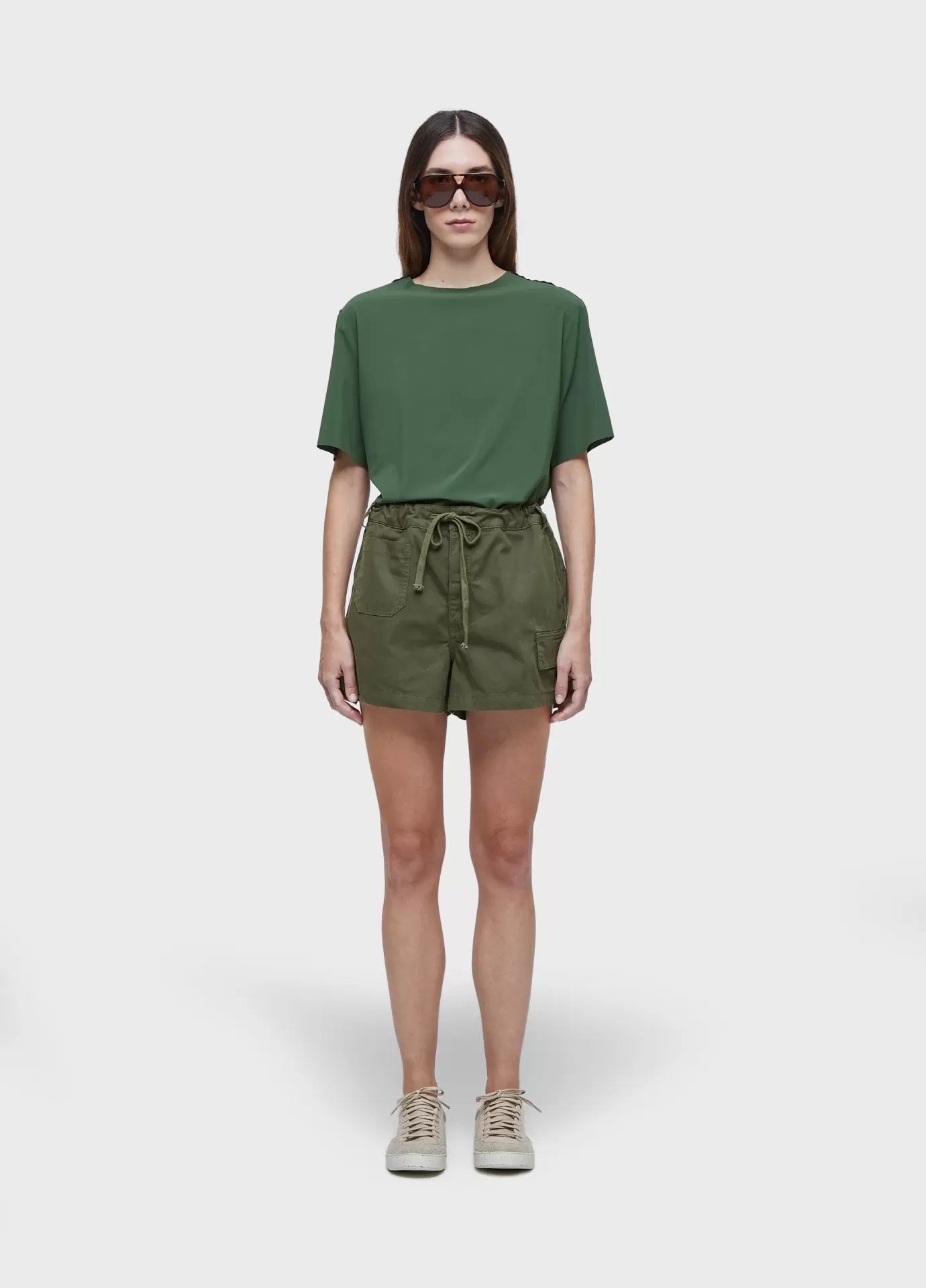 Short Bolsos Light Osklen Mulher com t-shirt verde escura, calções verde oliva, tênis bege e óculos de sol