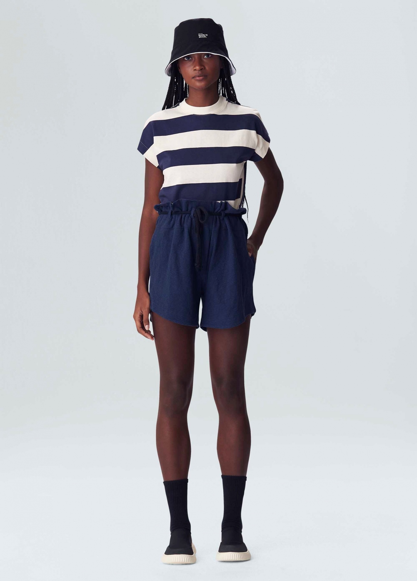 Short Clochard Node Osklen Mulher a usar t-shirt riscada branca e azul, calções azuis, chapéu preto, meias e ténis pretos