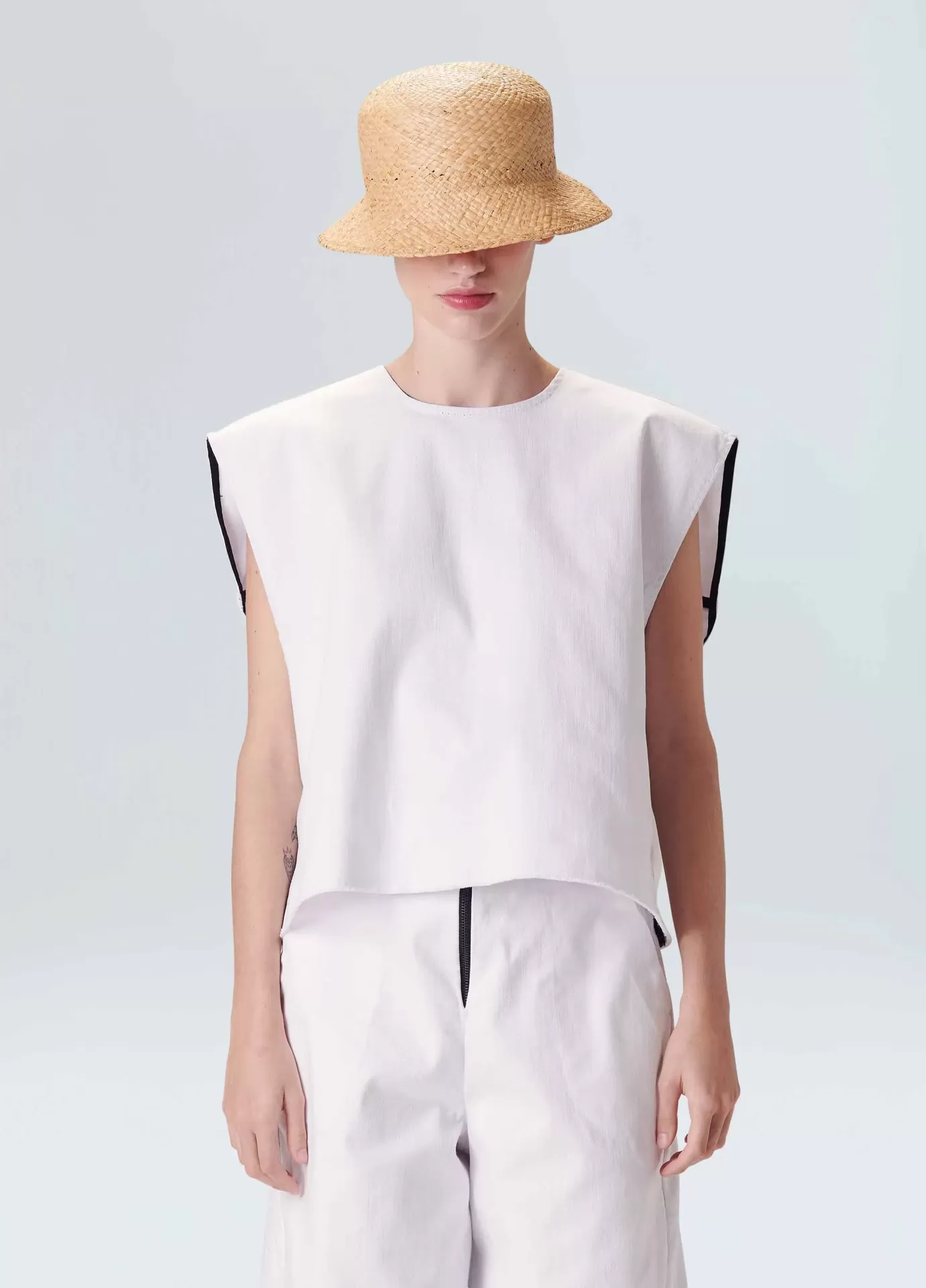 Colete Reversible Osklen Pessoa com chapéu de palha e roupa branca sobre fundo cinza claro