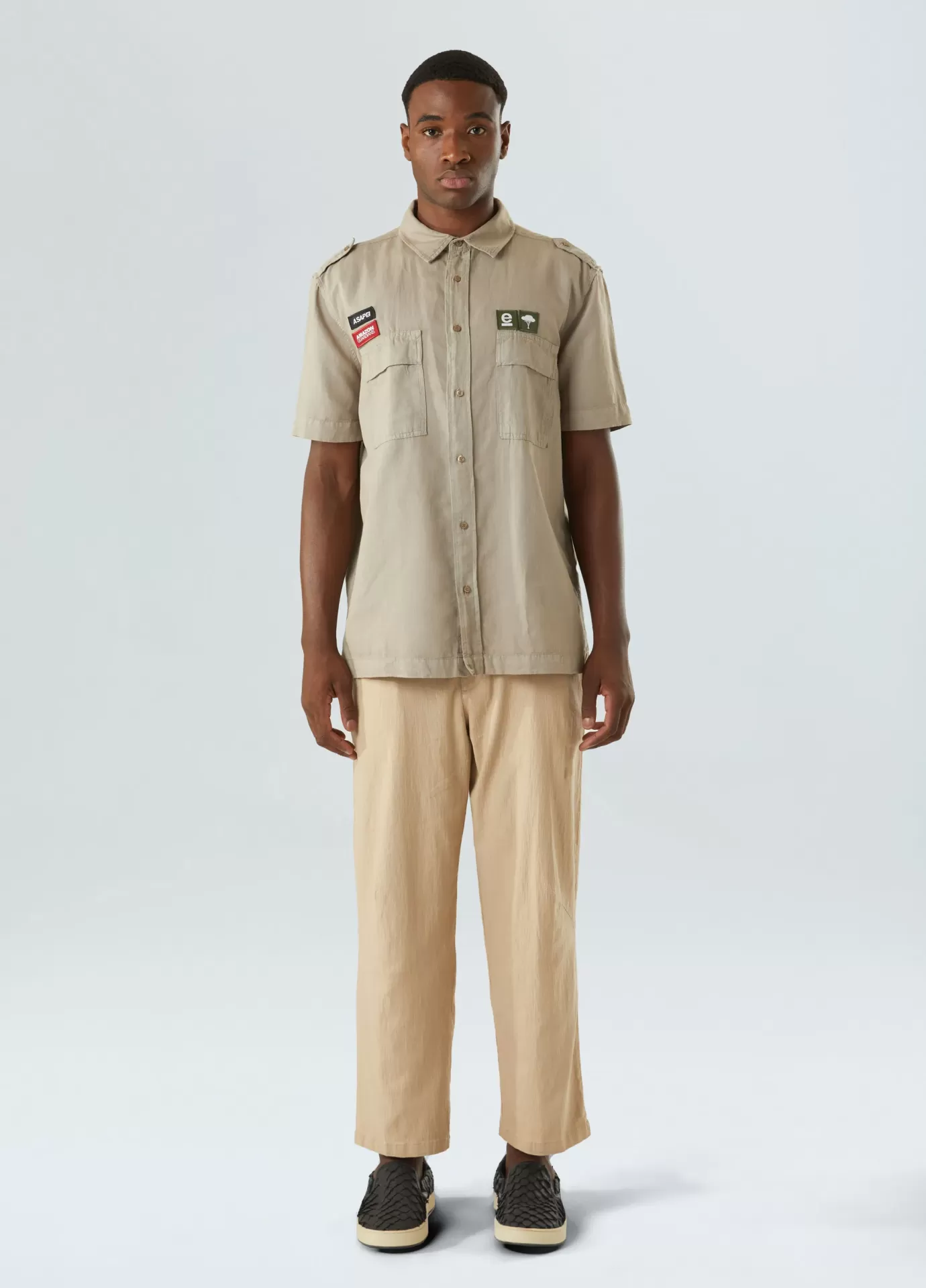 Camisa Utility Hemp Patch Osklen Homem com camisa bege de manga curta com etiquetas, calças largas bege e sapatos pretos em fundo cinza claro.