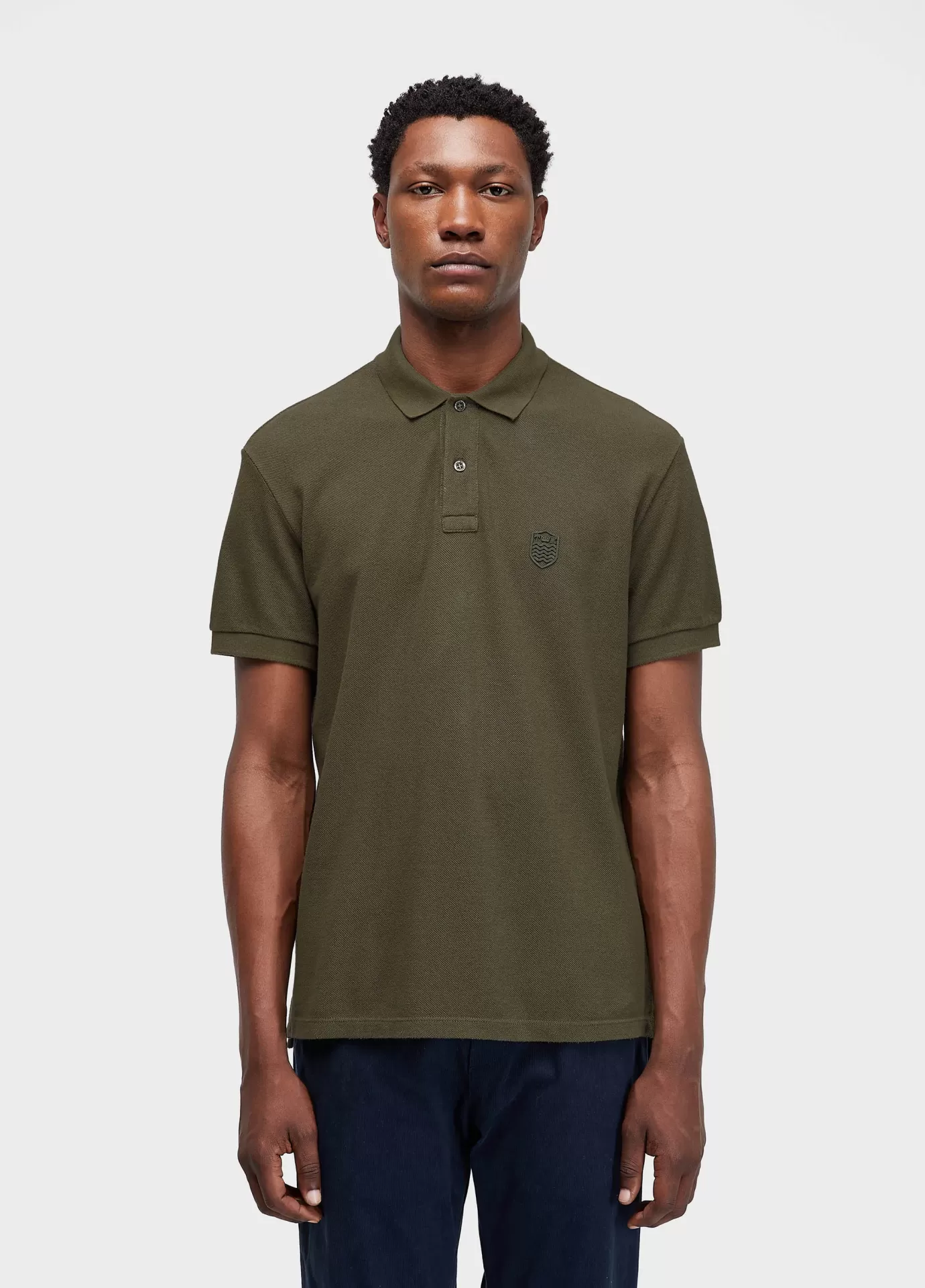 Polo Brasao Embossed Color Osklen Homem a vestir polo verde-oliva de manga curta com calças azuis, fundo branco
