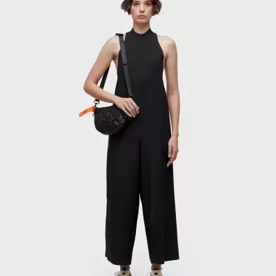 Mulher vestindo macacão preto sem mangas com gola alta, ténis brancos e mala preta com alça larga e detalhe laranja