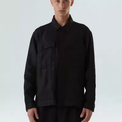 Homem com camisa preta de manga comprida e calças pretas em fundo cinzento claro