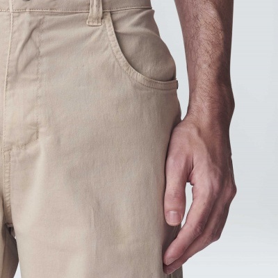 Calças bege com bolso lateral e botão prateado com braço ao lado em fundo branco