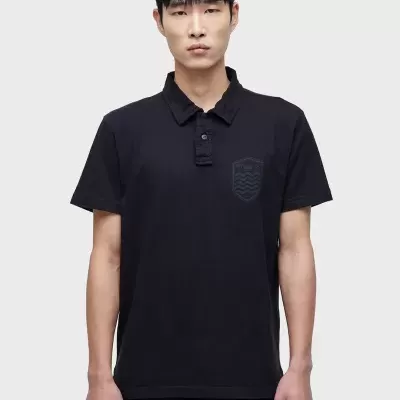 Polo preto de manga curta com emblema no peito usado por modelo masculino em fundo cinzento