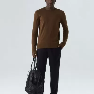 Homem com camisola castanha, calças pretas, sapatos claros e mala preta em fundo branco