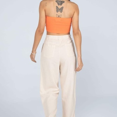 Mulher de costas com top laranja e calças beges largas e calçado preto numa parede cinza