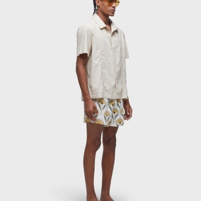 Homem com camisa bege de manga curta e calções florais, chinelos pretos e óculos de sol amarelos