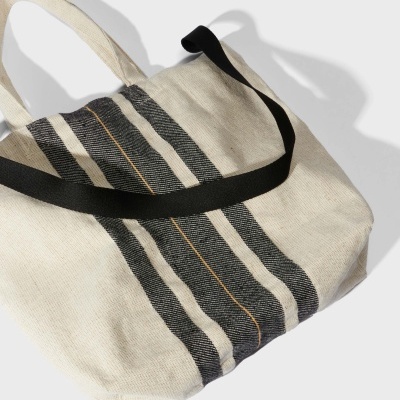 Mala de lona bege com riscas pretas verticais e linha laranja, alça preta.