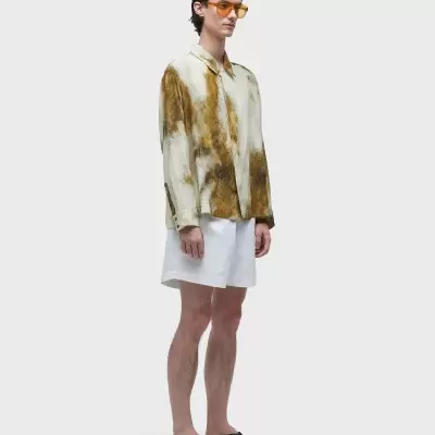 Homem com camisa branca manchada castanha, calções brancos e chinelos pretos