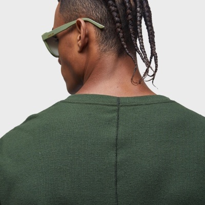 Pessoa de costas com cabelo trançado, óculos de sol verdes e camiseta verde escuro.