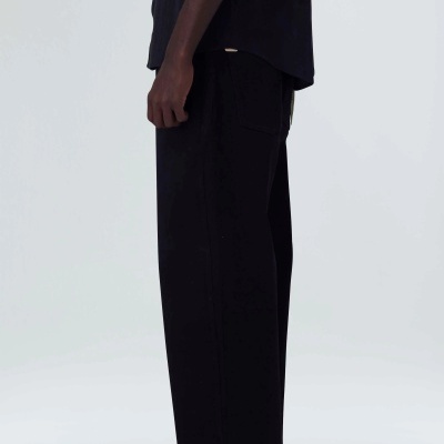 Pessoa de lado vestindo calças largas pretas com bolso traseiro, camisola preta e sapatos pretos num fundo branco.