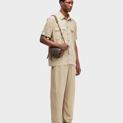 Homem com camisa bege, calças largas bege, sandálias castanhas e mala de ombro castanha com padrão texturizado.