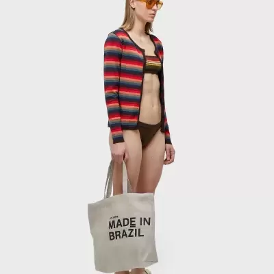 Mulher com casaco às riscas coloridas, biquíni e saco de lona com texto 'MADE IN BRAZIL'