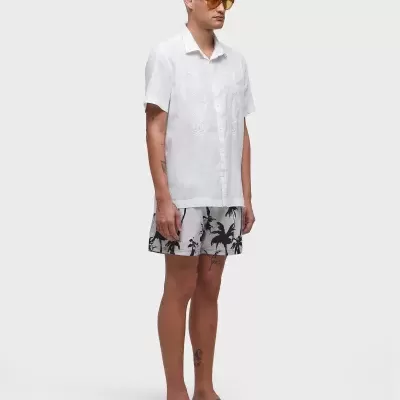 Homem vestido com camisa branca, calções estampados cinza e chinelos pretos, óculos de sol laranja