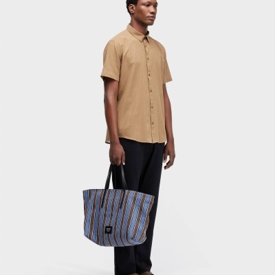 Homem com camisa bege de manga curta, calças pretas, sandálias castanhas e tote bag listrada azul, castanho e branco.