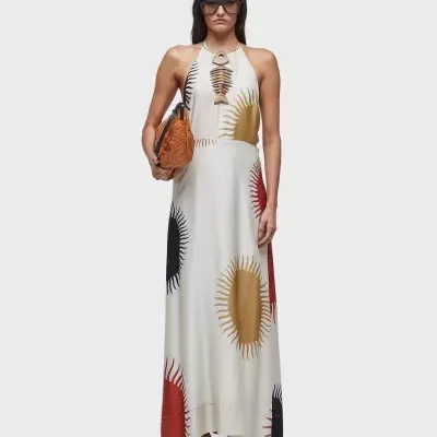 Modelo com vestido maxi branco estampado com formas solares, bolsa castanha, óculos escuros e sandálias.
