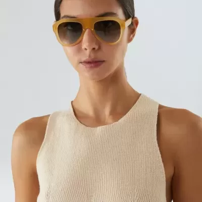 Óculos de sol com armação amarela e lentes escuras, modelo com vestido bege claro