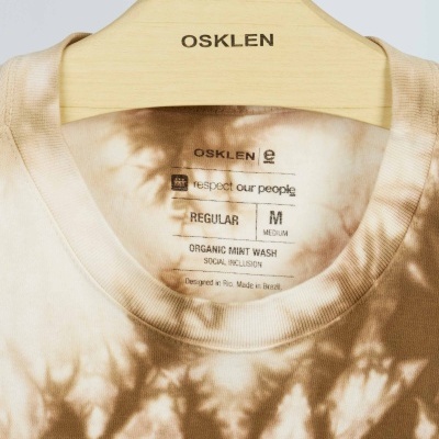 T-shirt tie-dye bege e castanha com etiqueta OSKLEN pendurada num cabide de madeira