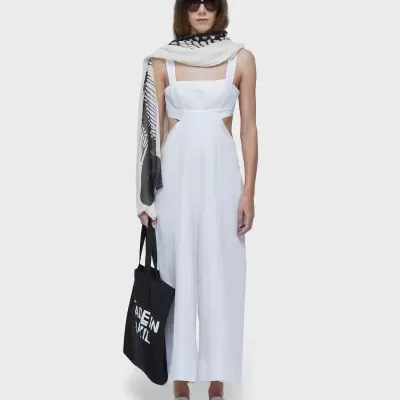 Modelo vestindo macacão branco, lenço bege e preto, óculos escuros, mochila preta com texto e sandálias brancas.