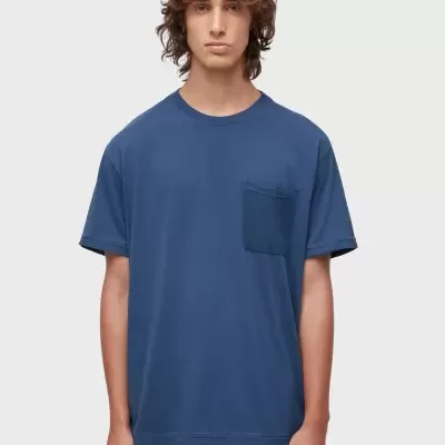 Homem com t-shirt azul e calças castanhas frente a fundo branco