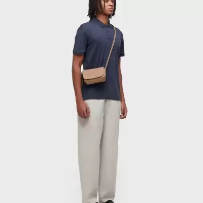 Homem com polo azul, calças bege, sapatos pretos e bolsa bege de tiracolo