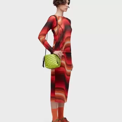 Mulher com vestido colorido de padrão ondulado, meias e sapatilhas, e mala verde de padrão escamas, em fundo cinza